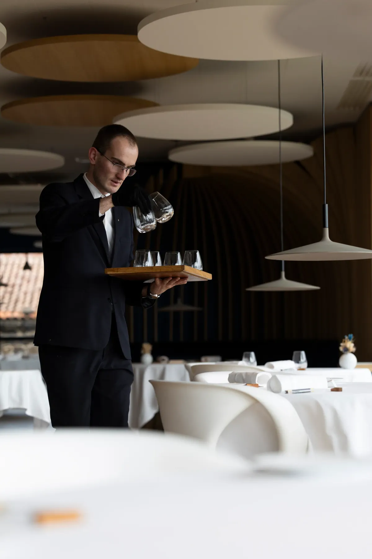 Un serveur en costume noir tient un plateau avec des verres à vin dans un restaurant élégant aux tables dressées