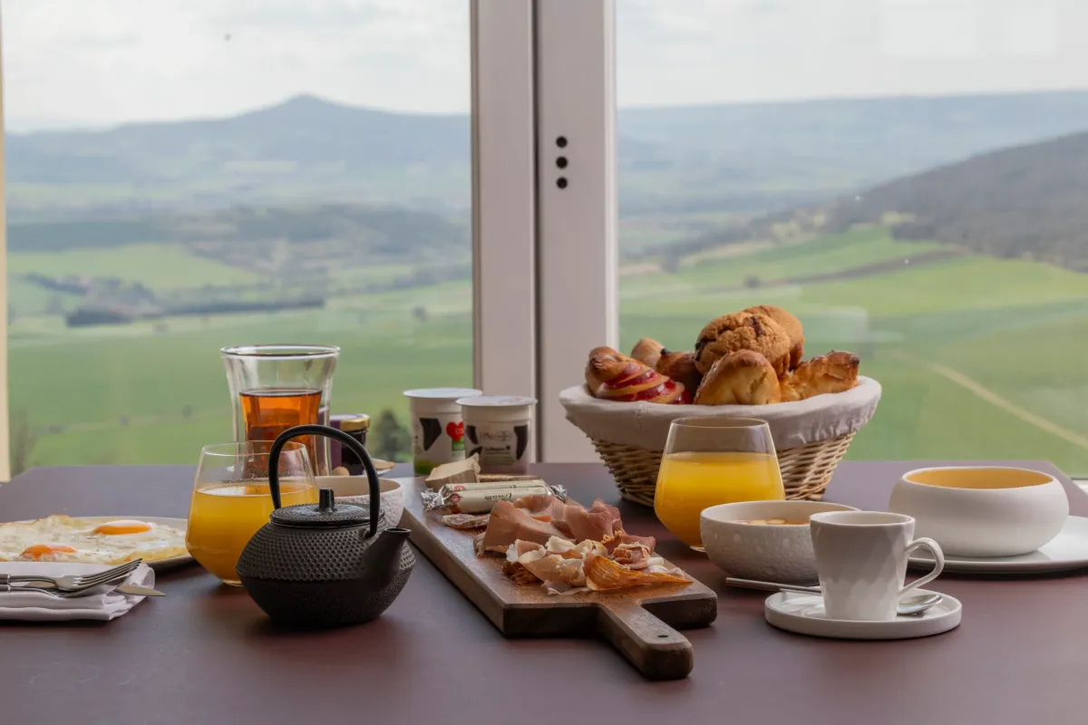 Table de petit-déjeuner avec œufs, bacon, pain, viennoiseries, jus d'orange, thé et yaourts devant une fenêtre offrant une vue sur des collines verdoyantes