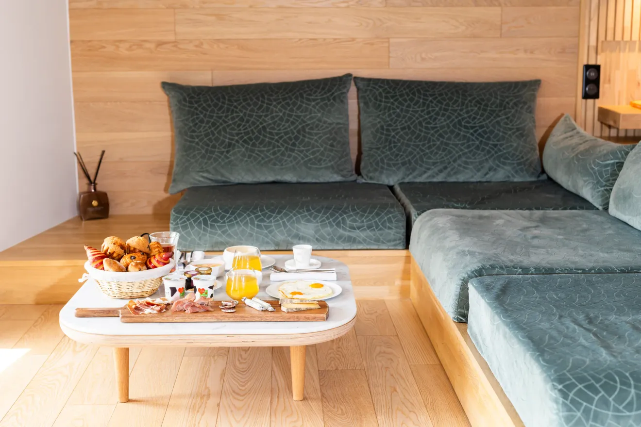 Un plateau de petit-déjeuner avec des œufs, des croissants, du jambon, du fromage, du jus d'orange et d'autres aliments est posé sur une table basse devant un canapé vert avec des coussins à motifs