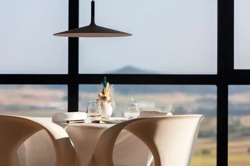 Table dressée avec verres, vase et nappe blanche devant une grande fenêtre offrant une vue sur un paysage montagneux