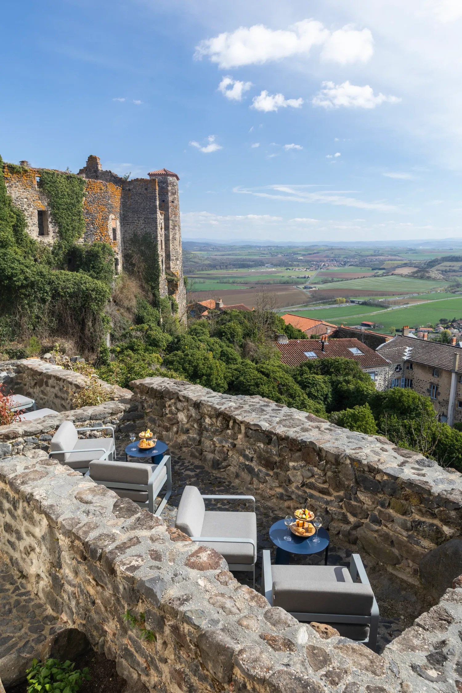 Vue depuis un château en ruine avec des canapés et tables basses sur une terrasse en pierre, surplombant une campagne vallonnée
