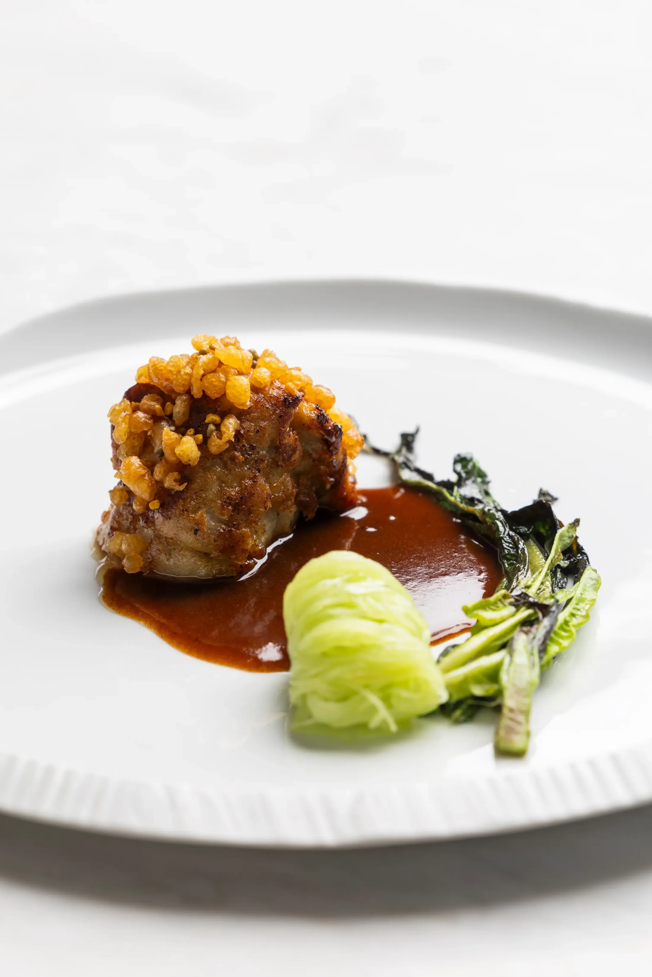Un plat composé d'une boule de viande croustillante, d'une purée verte et de légumes verts dans une sauce brune, présenté sur une assiette blanche