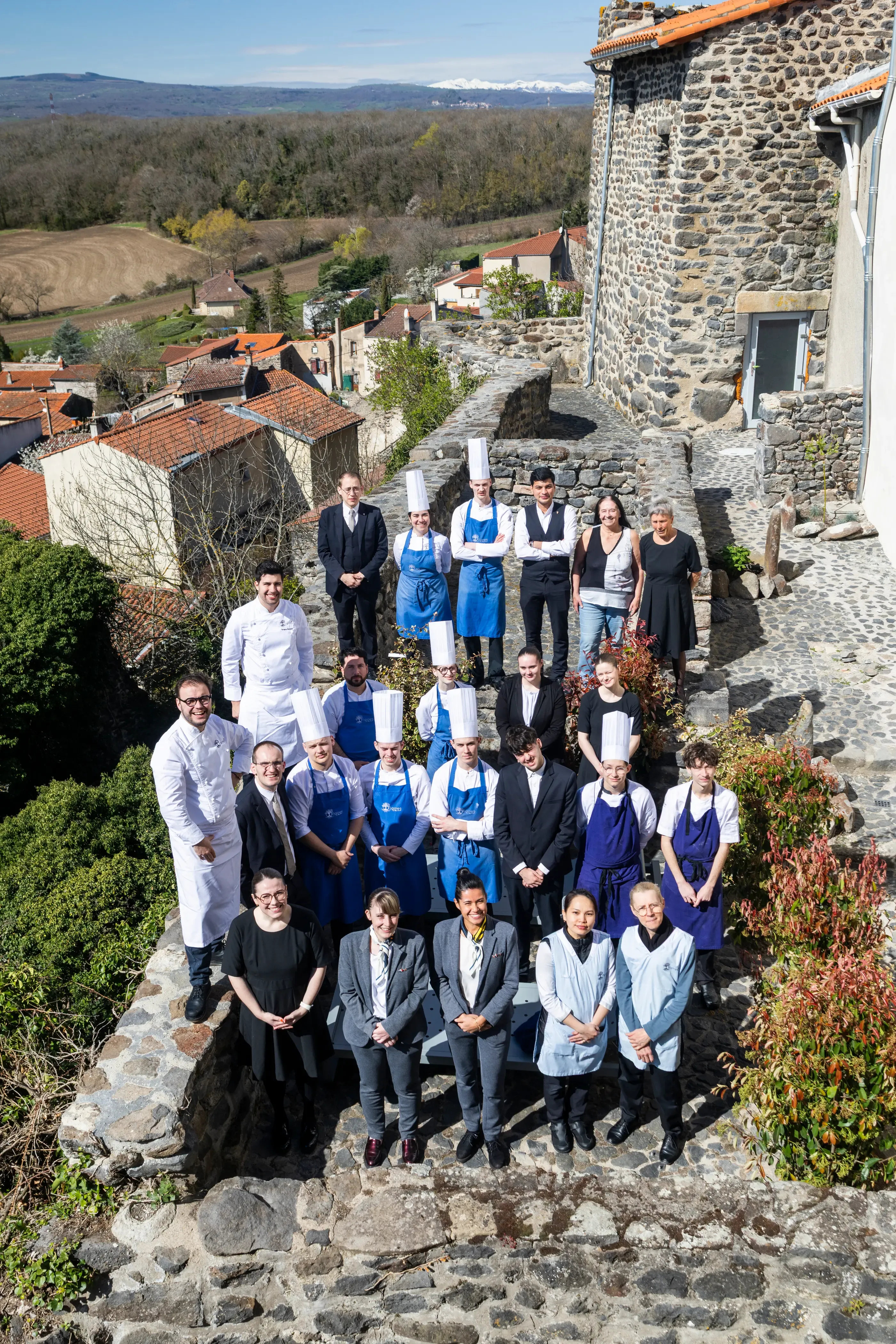 Un groupe de personnes en tenue de cuisine et en vêtements formels pose devant un bâtiment en pierre dans un village avec vue sur la campagne