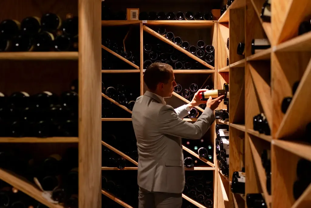 Un homme en costume place une bouteille de vin dans une cave à vin en bois remplie de bouteilles