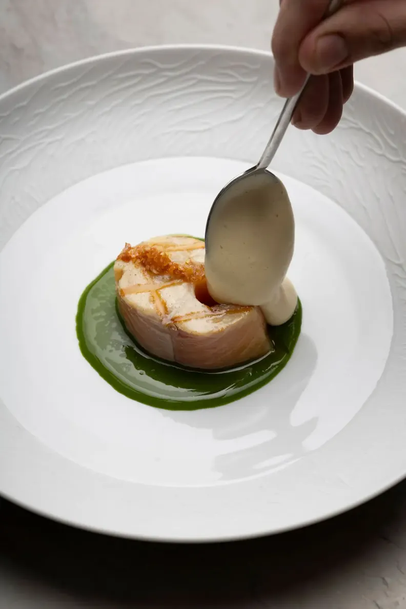 Un morceau de poisson grillé est posé sur une sauce verte, avec une sauce blanche ajoutée à la cuillère