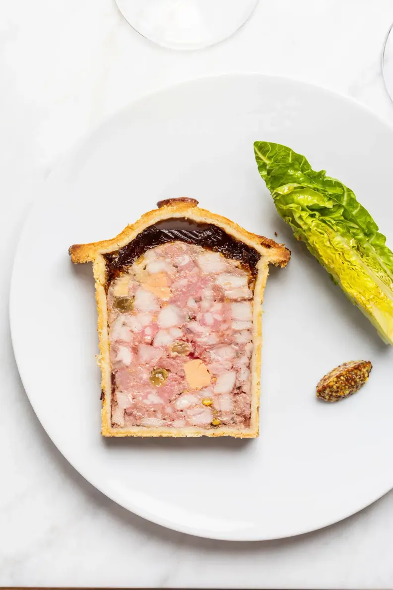 Une part de pâtisserie rectangulaire garnie de viande et de gelée, accompagnée d'une feuille de laitue et d'une graine sur une assiette blanche